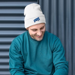 May include: A white beanie with the word "POP" embroidered in blue. The beanie is made of a soft, knitted material. The person in the image is wearing a teal sweatshirt.