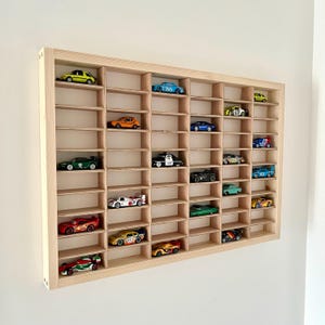 May include: A wooden display case with multiple compartments, showcasing a collection of colorful toy cars. The cars are various colors, including red, green, yellow, and blue. The case is made of light-colored wood and is mounted on a white wall.