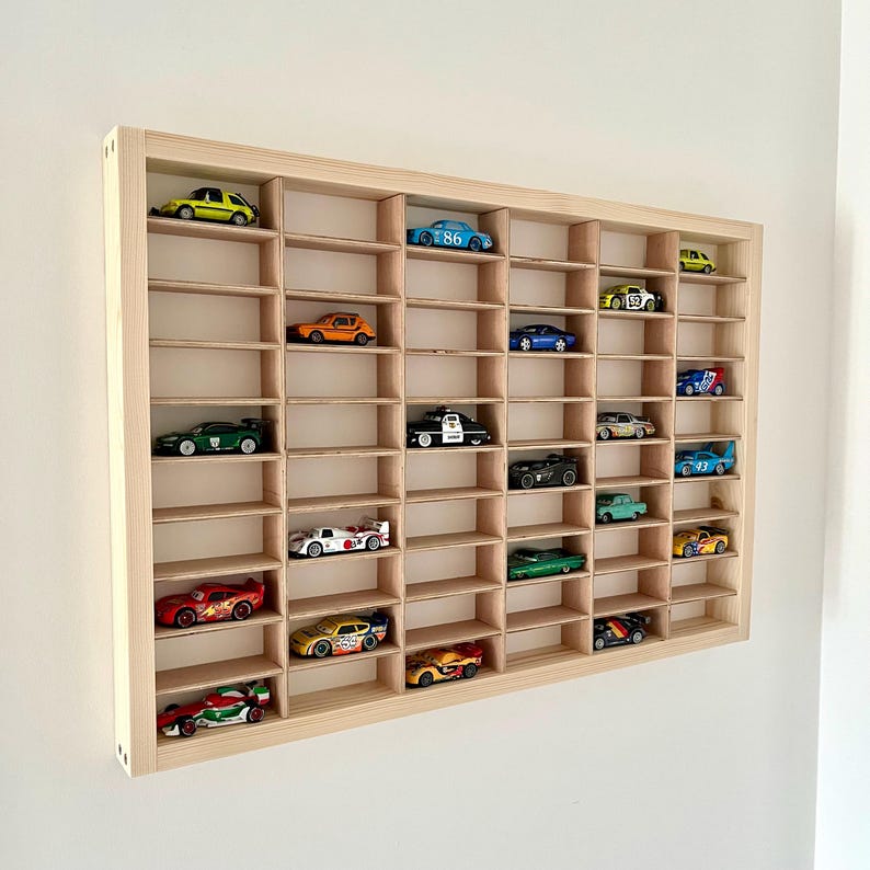 May include: A wooden display case with multiple compartments, showcasing a collection of colorful toy cars. The cars are various colors, including red, green, yellow, and blue. The case is made of light-colored wood and is mounted on a white wall.