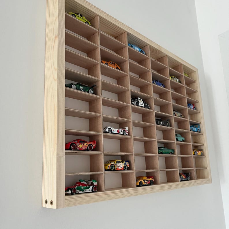 May include: A wooden display case with multiple compartments, filled with various toy cars in different colors. The case is mounted on a white wall, showcasing a collection of miniature vehicles.