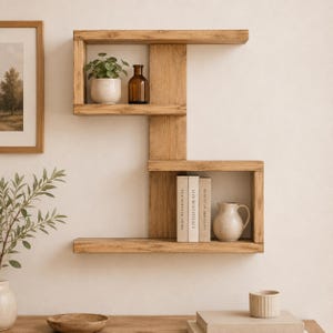 May include: A wooden wall shelf with a distinctive geometric design. It has three rectangular compartments, displaying a small potted plant, a brown glass bottle, books, and a ceramic jug. The natural wood grain gives a rustic feel.