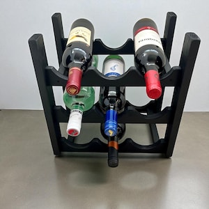 May include: A black wooden wine rack holding six bottles of wine. The bottles are various colors, including red, green, and clear. The rack is made of dark wood and has a simple, functional design.