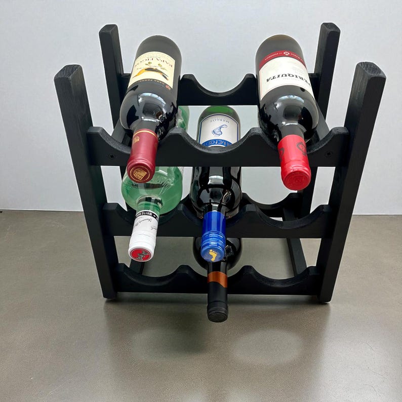 May include: A black wooden wine rack holding six bottles of wine. The bottles are various colors, including red, green, and clear. The rack is made of dark wood and has a simple, functional design.