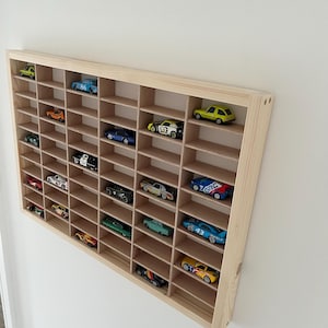 May include: A wooden display shelf with multiple compartments, filled with a collection of colorful toy cars. The shelf is made of light-colored wood and is mounted on a white wall. The toy cars are various colors and styles.