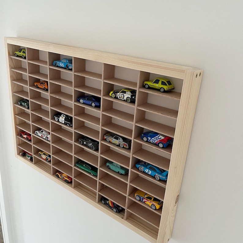 May include: A wooden display shelf with multiple compartments, filled with a collection of colorful toy cars. The shelf is made of light-colored wood and is mounted on a white wall. The toy cars are various colors and styles.