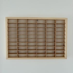 May include: A rectangular, light-colored wooden display case with multiple small, evenly spaced compartments. The case is mounted on a white wall. The compartments are designed to hold small collectibles or items.