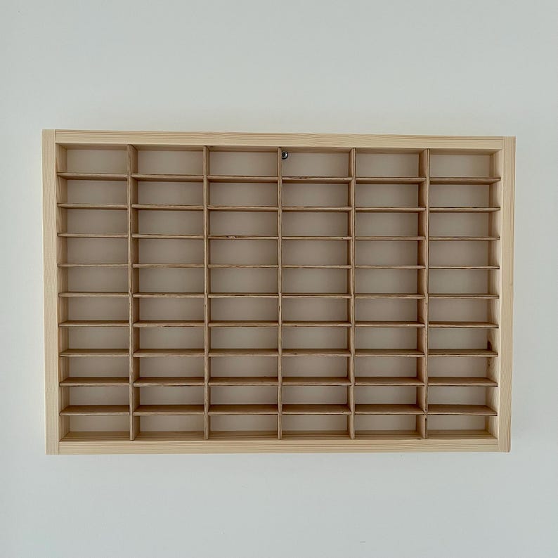 May include: A rectangular, light-colored wooden display case with multiple small, evenly spaced compartments. The case is mounted on a white wall. The compartments are designed to hold small collectibles or items.