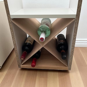 May include: A white and wood-toned wine rack with a geometric design, holding several bottles of wine and liquor. The bottles are various colors, including green, red, and black. The rack is on a light-colored wooden floor.