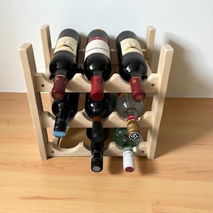 May include: A wooden wine rack holding several bottles of wine. The rack is made of light-colored wood and has a simple, open design. The wine bottles are various colors, with red wine bottles and a green bottle visible.
