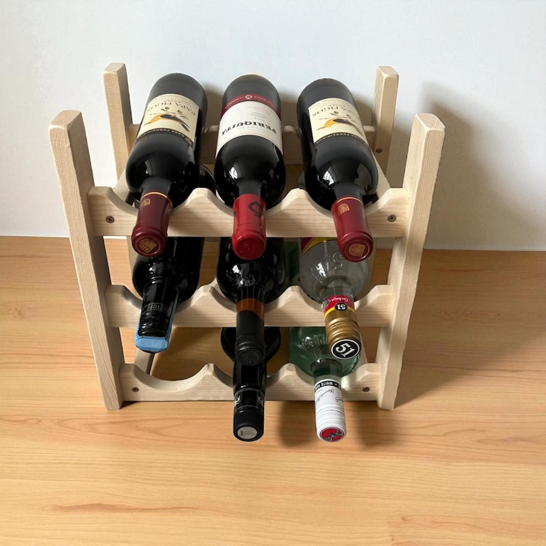 May include: A wooden wine rack holding several bottles of wine. The rack is made of light-colored wood and has a simple, open design. The wine bottles are various colors, with red wine bottles and a green bottle visible.