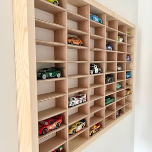 May include: A wooden display case with multiple cubby holes, each holding a toy car. The cars are various colors and styles, including race cars and classic models. The case is made of light-colored wood and mounted on a white wall.