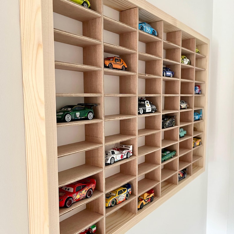 May include: A wooden display case with multiple cubby holes, each holding a toy car. The cars are various colors and styles, including race cars and classic models. The case is made of light-colored wood and mounted on a white wall.