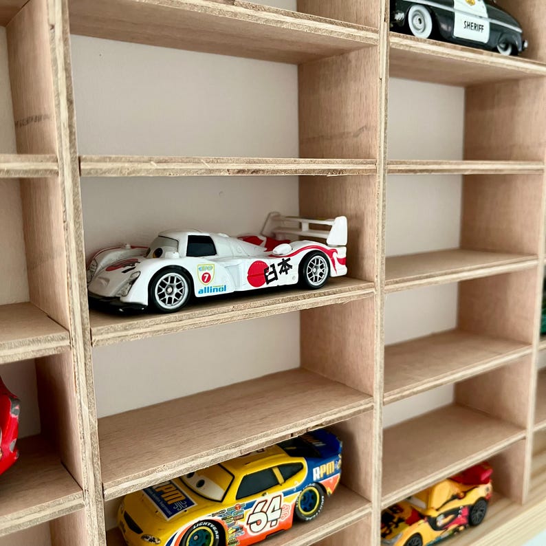 May include: A wooden display shelf with multiple compartments, each holding a toy car. The cars are various colors and designs, including a white race car with Japanese text and a yellow race car with the number 54.