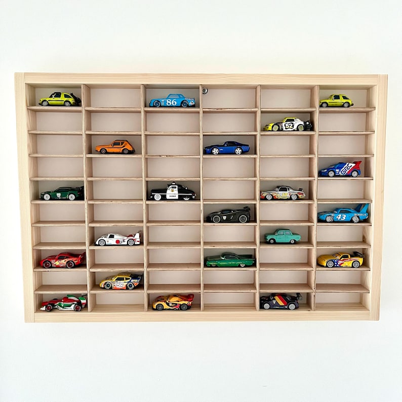 May include: A wooden display case with multiple compartments, filled with various toy cars in different colors and designs. The cars include a police car, race cars, and classic models. The case is made of light-colored wood.