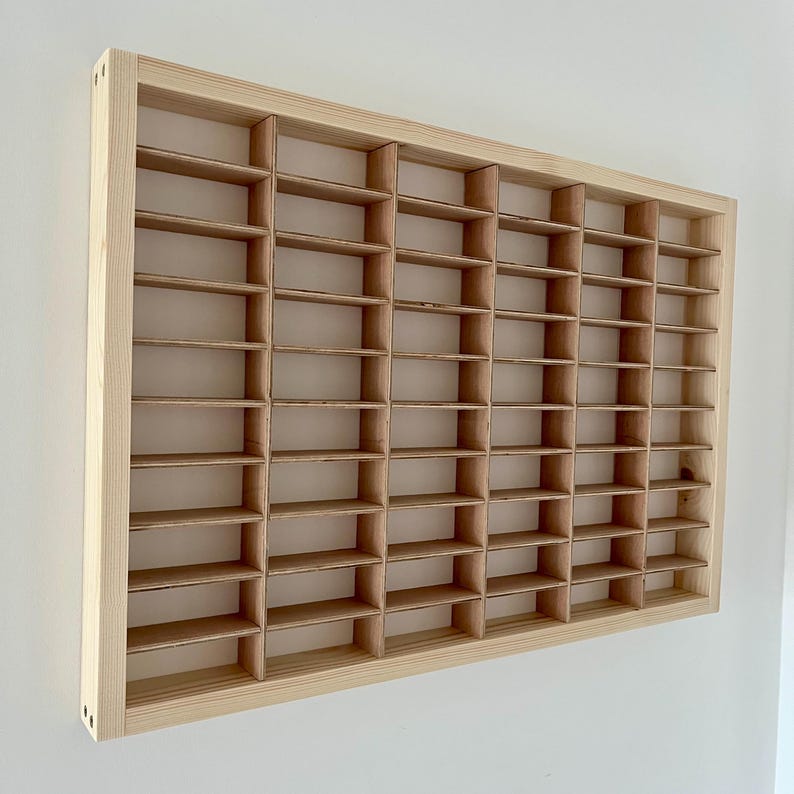 May include: A rectangular, light-colored wooden display shelf with numerous small, evenly spaced compartments. The shelf is mounted on a white wall, showcasing a grid-like structure ideal for organizing small items or collectibles. The natural wood grain is visible.