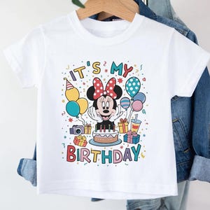 May include: White t-shirt featuring a colorful graphic of Minnie Mouse with a birthday cake, balloons, and the text "IT'S MY BIRTHDAY." The design includes a party hat, gifts, and confetti, creating a festive and celebratory theme.