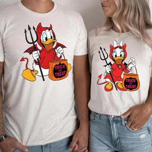 May include: Off-white t-shirts featuring cartoon characters dressed as devils for Halloween. One shirt has Donald Duck with a trident and a "Trick or Treat" bag. The other shirt has Daisy Duck with a trident and a "Trick or Treat" bag.