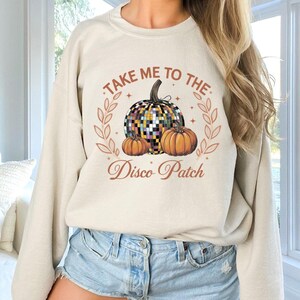 以下が含まれることがあります： クリーム色のスウェットシャツに、「Take me to the Disco Patch」という文字と、ディスコボールの南瓜と小さな南瓜2つのグラフィックがプリントされています。