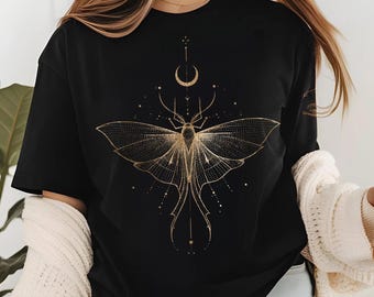 May include: Black t-shirt featuring a gold moth design with a crescent moon and celestial accents. The moth has detailed wings and a symmetrical pattern. The shirt is a casual style, suitable for everyday wear.