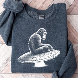 May include: Dark gray sweatshirt featuring a white illustration of a chimpanzee sitting on a UFO. The chimpanzee is facing left, with a detailed design. The sweatshirt is folded on a wooden surface.
