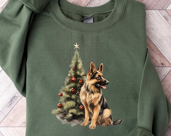 Op de afbeelding: Een groen sweatshirt met een kerstboom en een Duitse herder die op de voorkant zijn gedrukt. De hond zit naast de boom, die versierd is met rode kerstballen.