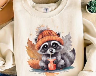 Pode incluir: Camisola creme com um guaxinim usando um gorro de malha laranja e bebendo de uma xícara. O guaxinim é cercado por folhas de outono e brilhos. A camisola é combinada com shorts jeans azuis e um chapéu marrom.