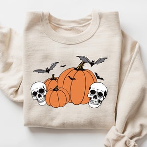 Könnte beinhalten: Ein beiger Pullover mit einem Halloween-Motiv mit drei orangefarbenen Kürbissen, zwei weißen Schädeln und drei schwarzen Fledermäusen.