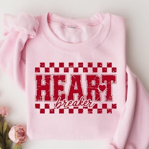 May include: Pink sweatshirt with a red checkered pattern and the text "HEART breaker" in a distressed font.