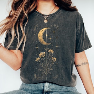 Puede incluir: Camiseta gris oscuro con un diseño de media luna dorada, estrellas y flores. La camiseta de manga corta está hecha de material suave y se combina con vaqueros azules y zapatillas blancas. El tema general es celestial y botánico.