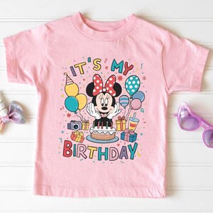 May include: Pink t-shirt with a colorful graphic featuring Minnie Mouse, balloons, a cake, and the text "IT'S MY BIRTHDAY." The design includes a camera, gifts, and a party hat. A pair of purple heart-shaped sunglasses is on the right.