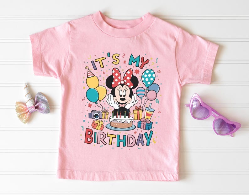 May include: Pink t-shirt with a colorful graphic featuring Minnie Mouse, balloons, a cake, and the text "IT'S MY BIRTHDAY." The design includes a camera, gifts, and a party hat. A pair of purple heart-shaped sunglasses is on the right.