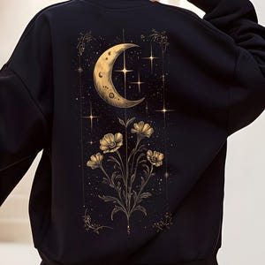 May include: Black sweatshirt featuring a celestial design. The back of the sweatshirt displays a gold crescent moon, stars, and floral elements. The design is detailed with gold accents, creating a striking contrast against the black fabric.