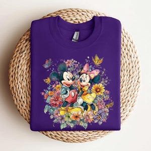 May include: A purple sweatshirt with a colourful illustration of Mickey and Minnie Mouse amidst flowers and butterflies. Mickey wears red shorts, and Minnie a yellow dress and pink bow. The design is a bright, whimsical illustration, perfect for Disney fans.