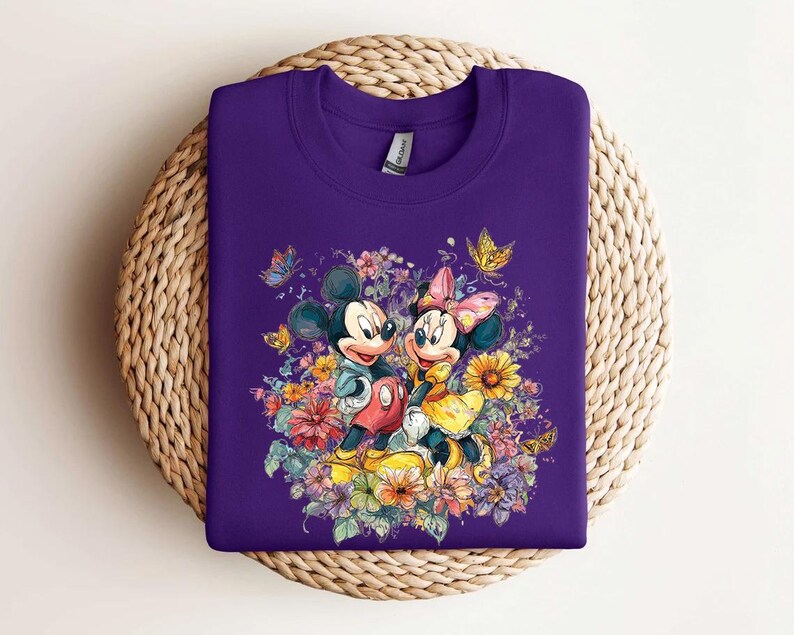May include: A purple sweatshirt with a colourful illustration of Mickey and Minnie Mouse amidst flowers and butterflies. Mickey wears red shorts, and Minnie a yellow dress and pink bow. The design is a bright, whimsical illustration, perfect for Disney fans.