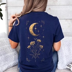 May include: Navy blue t-shirt featuring a gold illustration of a crescent moon, stars, and flowers. The celestial design is centered on the back of the shirt, creating a striking visual. The shirt is made of a soft, comfortable material.