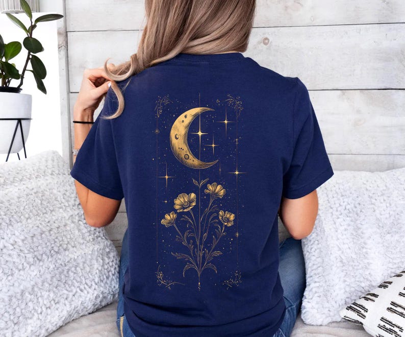 May include: Navy blue t-shirt featuring a gold illustration of a crescent moon, stars, and flowers. The celestial design is centered on the back of the shirt, creating a striking visual. The shirt is made of a soft, comfortable material.