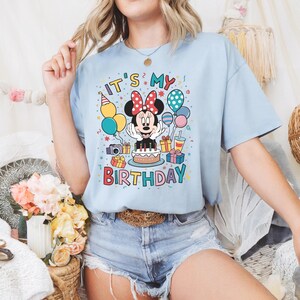 May include: Light blue t-shirt featuring a Minnie Mouse graphic with the text "IT'S MY BIRTHDAY". The design includes balloons, a birthday cake, and gift boxes. The shirt is made of a soft material.
