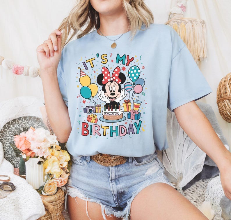 May include: Light blue t-shirt featuring a Minnie Mouse graphic with the text "IT'S MY BIRTHDAY". The design includes balloons, a birthday cake, and gift boxes. The shirt is made of a soft material.