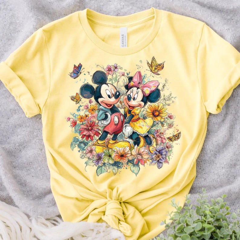 May include: Pale yellow t-shirt with a vibrant print of Mickey and Minnie Mouse amidst flowers and butterflies. Mickey is depicted in red shorts, Minnie in a yellow dress with a pink bow. The shirt is knotted at the waist, with rolled-up sleeves.