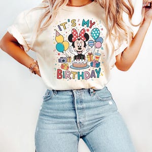 May include: Cream-colored t-shirt featuring a colorful Minnie Mouse birthday graphic. The design includes Minnie with a red bow, a cake, presents, balloons, and the text "IT'S MY BIRTHDAY". The shirt is paired with light blue jeans.