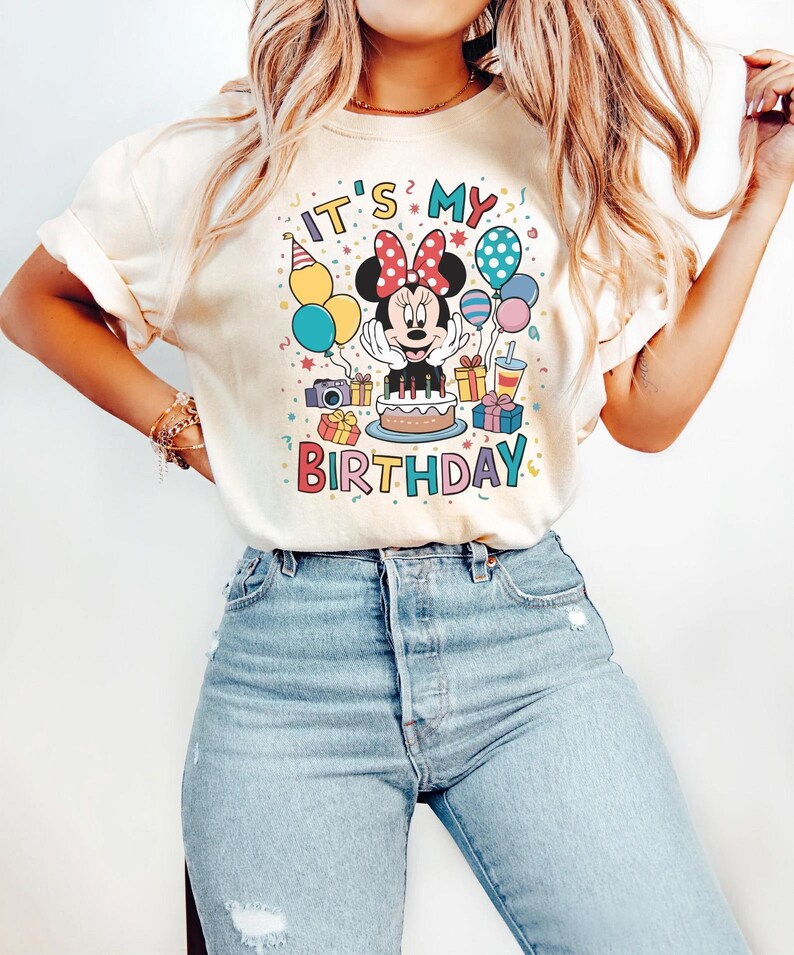May include: Cream-colored t-shirt featuring a colorful Minnie Mouse birthday graphic. The design includes Minnie with a red bow, a cake, presents, balloons, and the text "IT'S MY BIRTHDAY". The shirt is paired with light blue jeans.