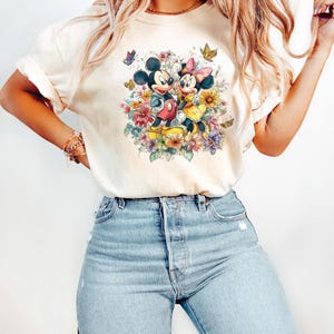 May include: Cream-coloured t-shirt featuring a colourful floral design with Mickey and Minnie Mouse. The cartoon characters are surrounded by flowers and butterflies. The shirt is paired with light blue denim jeans.