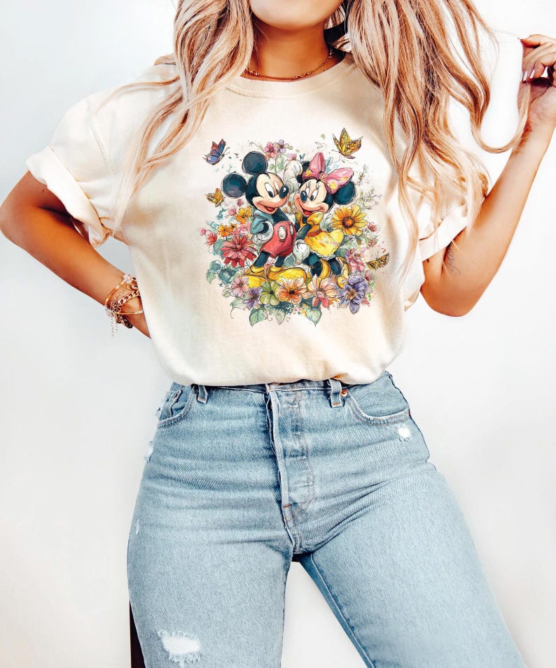 May include: Cream-coloured t-shirt featuring a colourful floral design with Mickey and Minnie Mouse. The cartoon characters are surrounded by flowers and butterflies. The shirt is paired with light blue denim jeans.