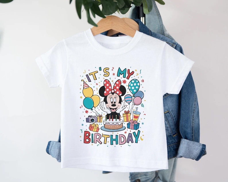 May include: White t-shirt featuring a colorful graphic of Minnie Mouse with a birthday cake, balloons, and the text "IT'S MY BIRTHDAY." The design includes a party hat, gifts, and confetti, creating a festive and celebratory theme.