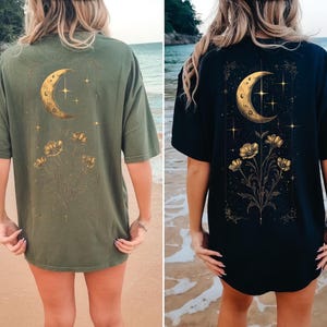 May include: Two t-shirts, one olive green and one navy blue, each featuring a gold crescent moon, stars, and floral design on the back. The shirts are short-sleeved and appear to be made of a soft material.