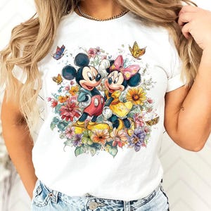 May include: White t-shirt with a colourful illustration of Mickey and Minnie Mouse amidst flowers and butterflies. Mickey is in red shorts and a blue jacket, Minnie in a yellow dress and pink bow. The design is bright and whimsical.