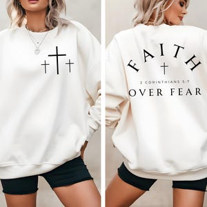Peut inclure: Un sweat-shirt blanc avec un motif de croix noire sur le devant et le texte "FAITH OVER FEAR 2 CORINTHIANS 5:7" au dos.