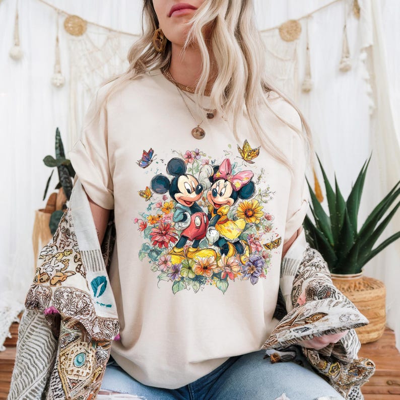 May include: Cream-coloured t-shirt featuring Mickey and Minnie Mouse surrounded by colourful flowers and butterflies. Mickey wears a blue jacket and red shorts, while Minnie dons a yellow dress and pink bow. The design is a whimsical, floral illustration.