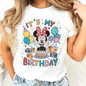 May include: White t-shirt featuring a colorful graphic of Minnie Mouse celebrating a birthday. The design includes the text "IT'S MY BIRTHDAY" with balloons, gifts, and a cake. The shirt is a classic crew neck style.