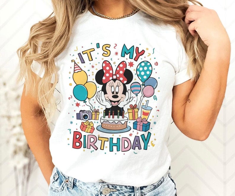 May include: White t-shirt featuring a colorful graphic of Minnie Mouse celebrating a birthday. The design includes the text "IT'S MY BIRTHDAY" with balloons, gifts, and a cake. The shirt is a classic crew neck style.